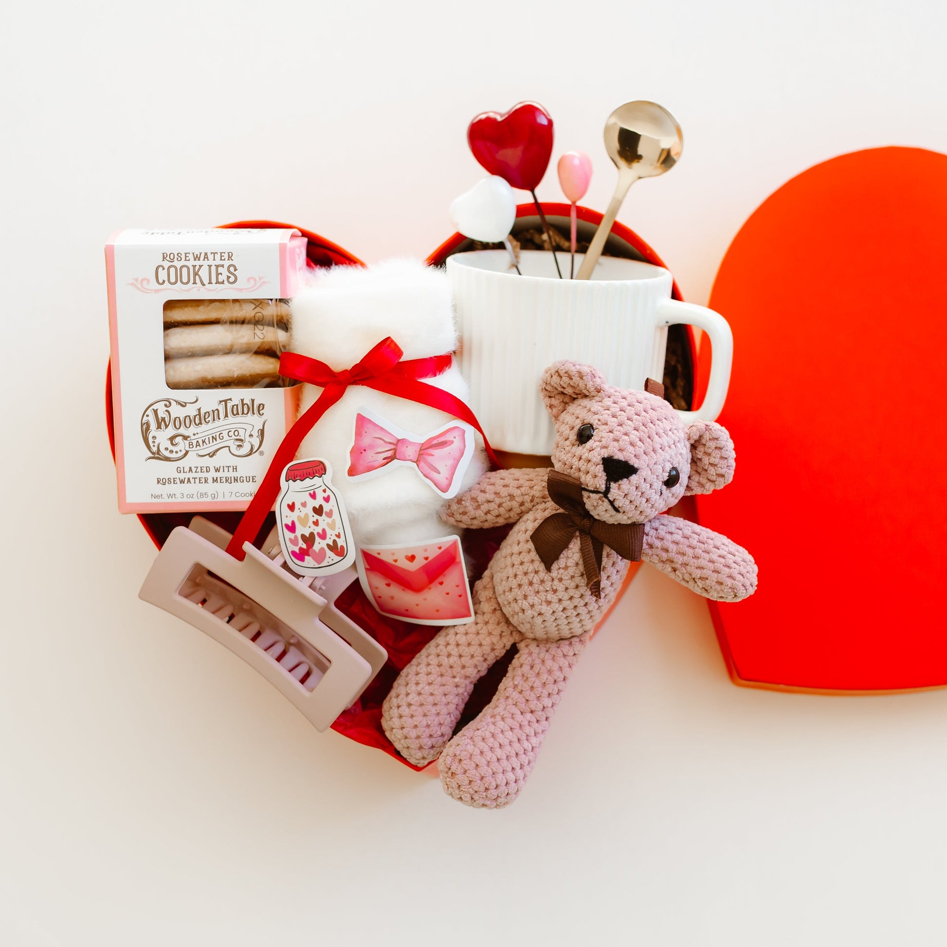Personalized Valentine’s Day heart-shaped gift box with teddy bear, rosewater cookies, socks, mug with gold spoon, and romantic decorations